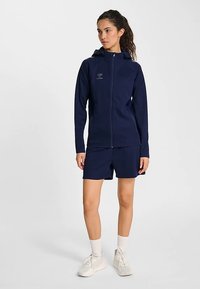 Marinblå sportjacka med huva och dragkedja, ihop med marinblå shorts. Har en slät textur och modern passform.
