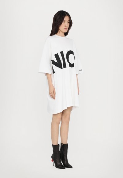 Biele oversized tričko šaty s tučným čiernym logom "NIC". Krátke rukávy, vysoký golier a mierne nepravidelný lem. Skombinované s čiernymi členkovými čižmami.