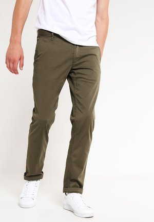 Pantaloni di cotone verde oliva con vestibilità slim, tasche anteriori e risvolti, abbinati a scarpe da ginnastica bianche e una maglietta bianca semplice.