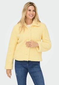 Yellow textured fleece jacket with a collar, button closure, and long sleeves. Worn over blue jeans in a neutral background.