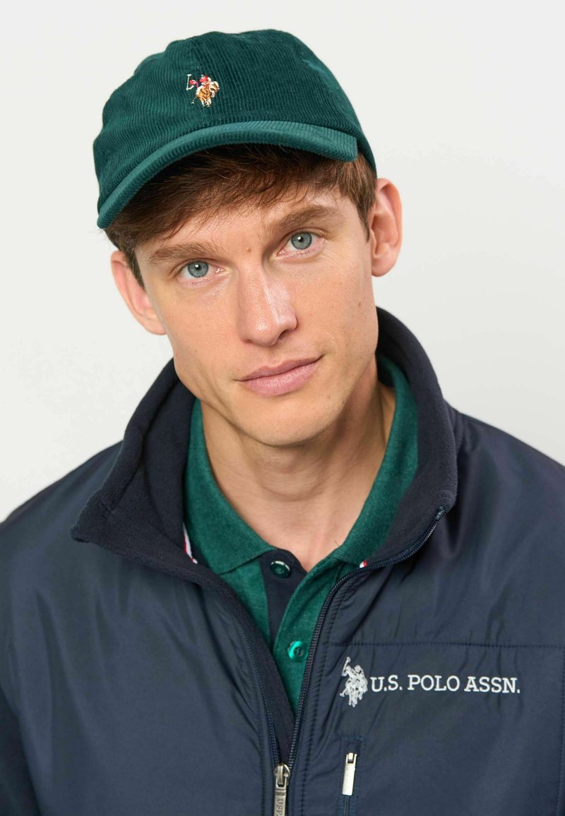Jeune homme aux yeux bleus, portant une casquette verte et un polo, ainsi qu'une veste bleu marine avec le logo U.S. Polo Assn., regarde doucement la caméra.