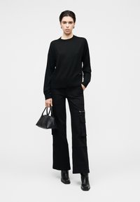 Svart strikket genser med rund hals, kombinert med løse svarte cargobukser med lommer. Modellen holder en liten svart håndveske.