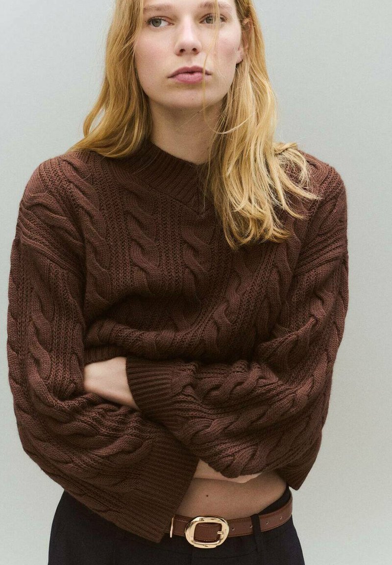 Brauner Kabelstrickpullover mit langen Ärmeln und lockerer Passform, mit tiefem V-Ausschnitt und gerippten Bündchen. Getragen mit einem braunen Gürtel.