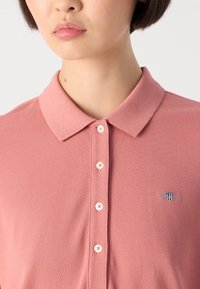 Detailní záběr na osobu v prachově růžovém polo tričku na knoflíky s límečkem a malým vyšitým logem na hrudi.