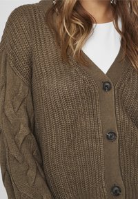 Brun chunky stickad cardigan med stora svarta knappar, bärs över en vit topp, med flätad kabelstickning på ärmarna.