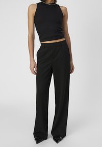 Slanke persoon in een mouwloze zwarte top, hooggewaist brede zwarte broek, puntige zwarte schoenen en een zilveren kettinghalsketting staat tegen een grijze achtergrond.