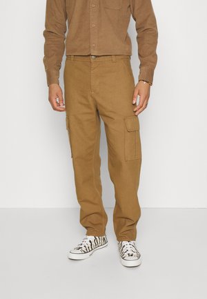 Pantaloni cargo marroni con due tasche laterali a toppa, realizzati in tessuto robusto, abbinati a scarpe da ginnastica a fantasia bianco e nero.