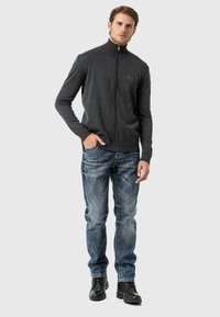 Grauer Zip-Up-Pullover mit hohem Kragen, kombiniert mit hellblauen Jeans und schwarzen Stiefeletten. Der Pullover zeichnet sich durch eine gerippte Struktur und eine schlanke Passform aus.