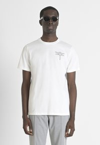 Camiseta de algodón blanca con un gráfico de libélula en el pecho. Mangas cortas, cuello redondo y un corte relajado. Combinada con pantalones grises.