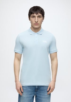 Jeune homme portant un polo bleu clair Karl Lagerfeld et un jean bleu, debout devant un fond blanc uni.