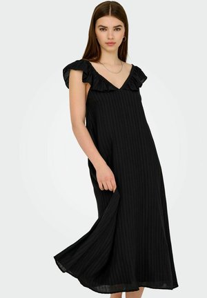 Femme jeune portant une robe midi sans manches noire avec un décolleté en V froncé, tenant l'ourlet de la robe contre un fond clair.