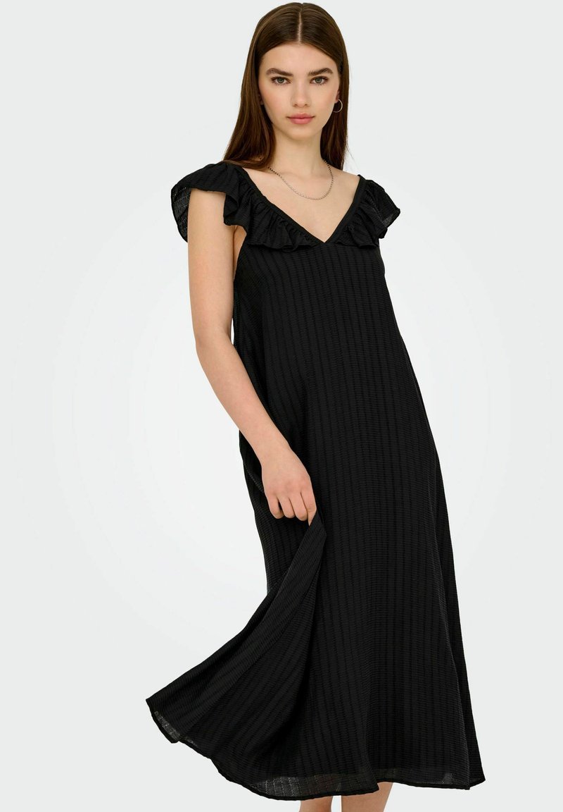 Joven mujer vistiendo un vestido midi sin mangas negro con un escote en V con volantes, sosteniendo el dobladillo del vestido contra un fondo claro y liso.