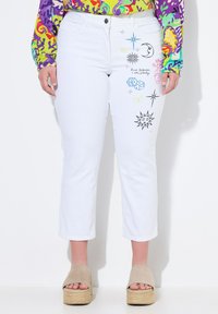 Valkoiset farkut, joissa on mustia, sinisiä ja pastellivärisiä brodeerattuja kuvioita, joissa on taivaallisia ja leikkisiä teemoja edessä.