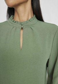 Grüne Bluse aus strukturiertem Stoff, hochgeraffter Ausschnitt, Schlüssellochöffnung und Knopfverschluss am Kragen. Lange Ärmel.