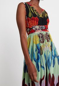 Robe longue à col en V, composée d'un corsage noir orné de motifs rouges et floraux, et d'une jupe verte décorée d'imprimés floraux colorés.