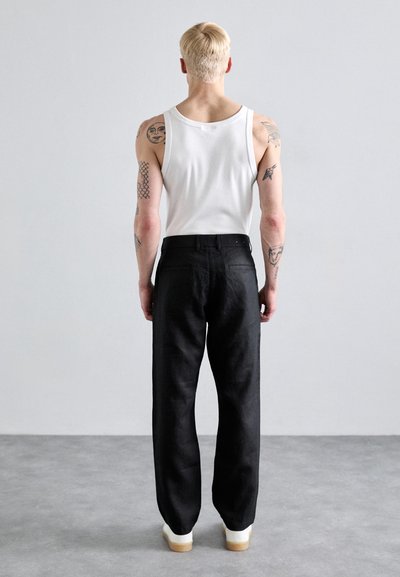 Osoba se světlými vlasy stojí čelem k jednoduché šedé zdi, má na sobě bílý nátělník, černé kalhoty, bílé boty a na obou pažích viditelné tetování.