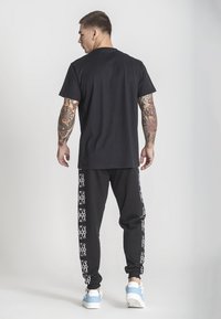 Svart t-shirt och svarta sweatpants med vita logotypmönster längs sidorna; modell bär vita och blå sneakers, vilket visar upp en avslappnad outfit.