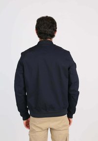 Homme avec des cheveux bruns portant une veste noire et un pantalon cargo beige, se tenant de dos devant un fond blanc uni.