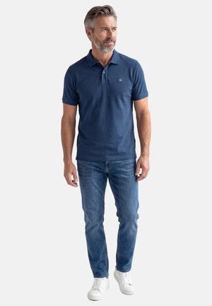 Mann mit grauem Haar und Bart, der ein marineblaues Polohemd, blaue Jeans und weiße Turnschuhe trägt und vor weißem Hintergrund steht.