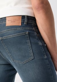 Sötétkék farmernadrág, testhezálló kialakítással. Bőr címkével ellátott, melyen a "Marc O'Polo Denim" felirat szerepel, valamint klasszikus hátsó zsebekkel rendelkezik.