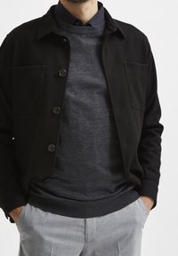 Svart knäppjacka med två bröstfickor över en mörkgrå tröja. Utrustningen kompletteras med ljusgrå skräddarsydda byxor.