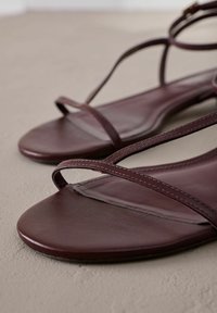 Sandalias de cuero burdeos oscuro con tiras delgadas entrecruzadas y suela plana. Textura suave con un detalle de costura mínimo.