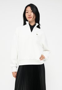 Giovane donna che indossa un felpa bianca con zip a quarti e un logo verde, accompagnata da una gonna plissettata nera, in piedi con una mano su un fianco.