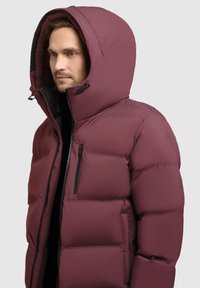 Mørkerød pufferjakke med en stor hætte, lynlåslomme på brystet og justerbare snore. Har quiltet tekstur og mellemlang design.