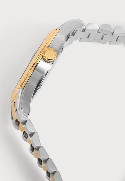 Vue de côté d'une montre-bracelet argentée et dorée avec une couronne en or et un bracelet à maillons métalliques sur un fond clair uni.