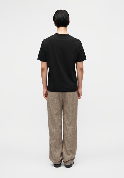 Μαύρο βαμβακερό μπλουζάκι με στρογγυλή λαιμόκοψη, συνοδευόμενο από ανοιχτό καφέ υφασμένα μάλλινα παντελόνια. Η όψη δείχνει την πίσω πλευρά της εμφάνισης.