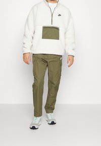 Valkoinen fleece-pullover vihreällä vetoketjutaskulla, yhdistetty oliivinvihreisiin cargo-housuihin ja sinivärisillä yksityiskohdilla varustettuihin lenkkitossuihin.