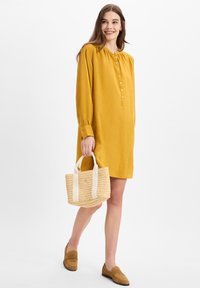 Sininen napillinen mekko, jossa on puhvihihat, yhdistettynä kudottuun olkilaukkuun ja vaaleanruskeisiin loafereihin, tasaisen taustan edessä.