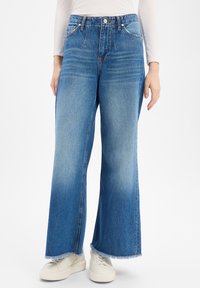 Hellblaue Jeans mit weitem Bein, verwaschenem Finish und ausgefranstem Saum. Mit fünf Taschen und einem Bund mit Knopfverschluss. Kombiniert mit weißen Turnschuhen.