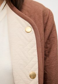 Vadderad jacka med en brun och krämfärgad färgskala, guldaccenter, en texturerad mönstring och en kontrasterande sydd kantdetalj.