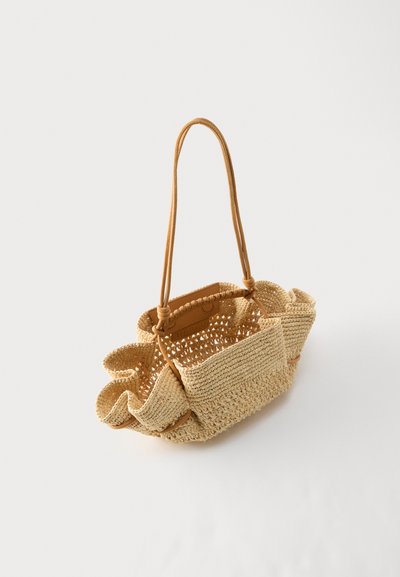 Μπεζ χρωματισμένη τσάντα πλεκτή με μακριές διπλές λαβές και βολάν στις πλευρές, εμφανίζεται σε απλό λευκό φόντο.