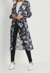 Žena v průhledném námořnickém květinovém dlouhém kardiganu, bílém tričku, černých upnutých džínách a bílých lodičkách na klínku se stříbrnou sponou.