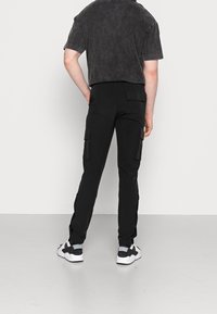 Person som står med ryggen mot kameran, iförd svarta cargo-byxor med sidofickor, en tvättad svart t-shirt med kort ärm och svart-vita sneakers.