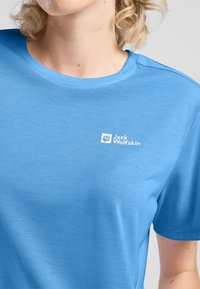 Lichtblauwe T-shirt met korte mouwen en een ronde hals, met het "Jack Wolfskin"-logo in het wit gedrukt op de bovenste linkerzijde. Glad van textuur.