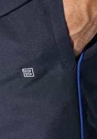 Dunkelblauer Stoff mit glatter Textur und dezentem Glanz, ausgestattet mit einem kleinen weißen Logo und einem kontrastierenden blauen Akzent an der Seitennaht.