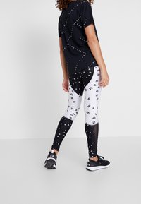 Svart T-shirt med vit cirkulär text, tillsammans med högmidjade leggings med ett djärvt svart- och vitt mönster. Svarta träningsskor.