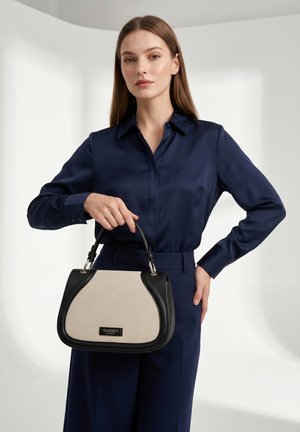 Donna con abbigliamento blu navy che tiene una borsa Diana&Co beige e nera con manici superiori su sfondo bianco uniforme.