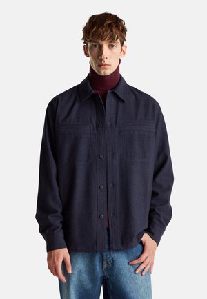 Dunkelblazer aus Wollmischung mit Kragen, Knopfverschluss, zwei Brusttaschen und langen Ärmeln, getragen über einem burgunderfarbenen Rollkragenpullover und Jeans.