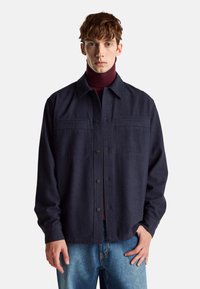 Chaqueta de mezcla de lana en color navy con cuello, cierre de botones, dos bolsillos en el pecho y mangas largas, llevada sobre un suéter de cuello alto burdeos y jeans de mezclilla.