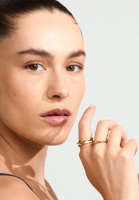 Gouden gedraaide ringen gedragen op een hand met een neutrale huidskleur. De achtergrond is lichtgrijs, wat de reflecterende textuur en het ontwerp van de ringen benadrukt.