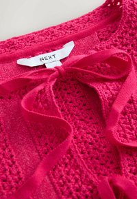 Cardigan tricotat croșetat într-o nuanță roz aprins, prevăzut cu un sistem de închidere prin legare și cu o etichetă inscripționată "NEXT" aproape de guler. Textură moale și design cu țesătură deschisă.