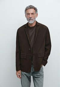 Blazer in camoscio marrone con due tasche frontali e due bottoni, abbinato a una camicia marrone e jeans in denim chiaro. Tessuto texturizzato, vestibilità sartoriale.