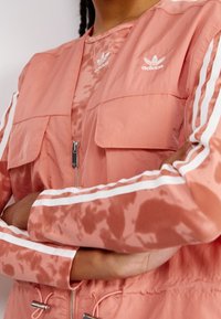 Person som bär en rosa Adidas-jacka med vita ränder på ärmarna och logotyp på bröstet, med armarna i kors, vilket visar blixtlås och framfickor.