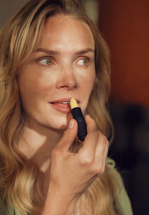 Schwarzer Lippenstift mit gelber Spitze, nahe den Lippen gehalten. Natürlicher Hautton und welliges, blondes Haar in einer sanft beleuchteten Umgebung.