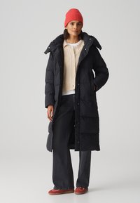 Doudoune noire avec capuche, entièrement zippée, associée à un cardigan beige, un jean foncé et des chaussures slip-on rouges. Tissu lisse, manches longues.