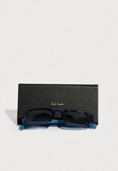 Čierne kožené púzdro na okuliare so zlatým logom „Paul Smith“, drží tmavé slnečné okuliare s priehľadnými modrými stranicami a obdĺžnikovými sklami.